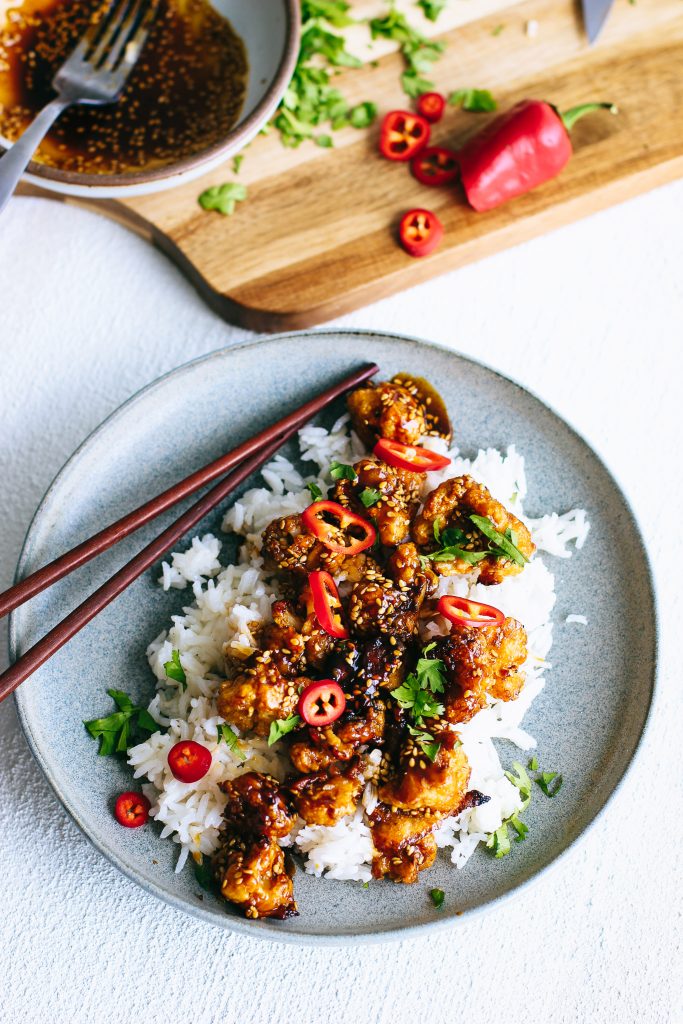 crispy cauliflower with red peppers and herbs over white rice with chopsticks on grey plate
