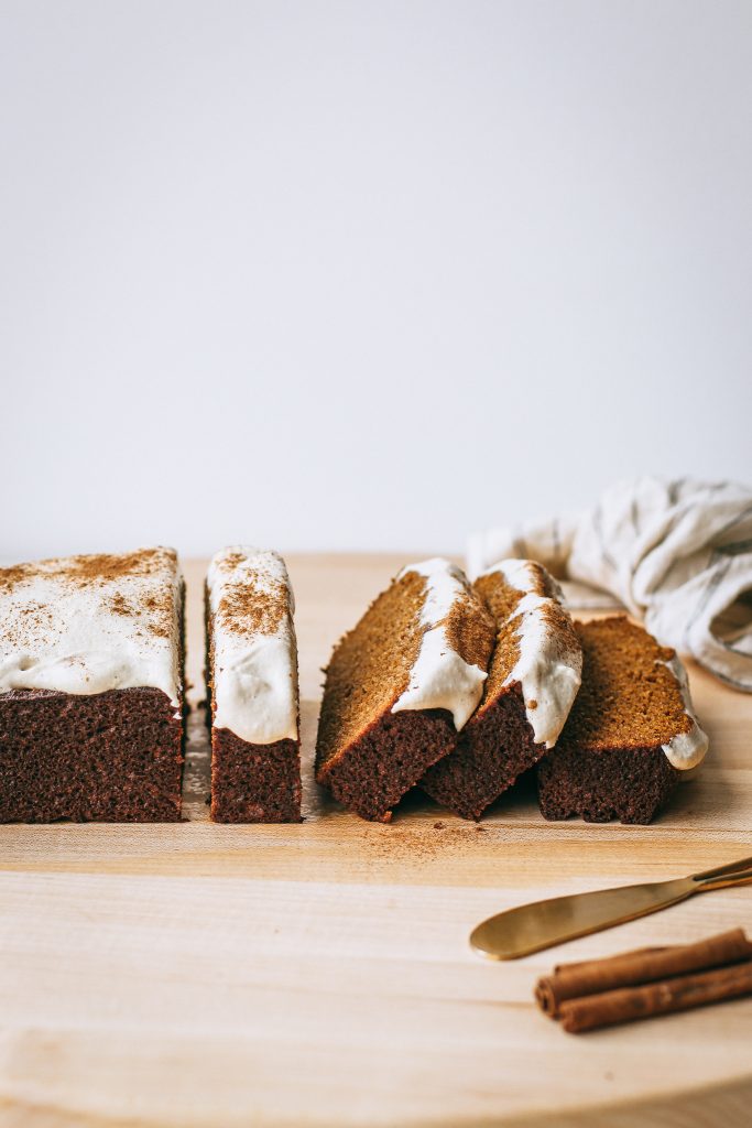 Pumpkin bread with whipped frosting and cinnamon topping on a wooden board with cinnamon sticks and a gold knife 
