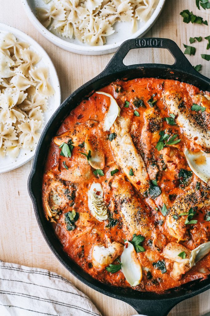 chicken and artichokes in creamy tomato sauce in skillet with plates of pasta