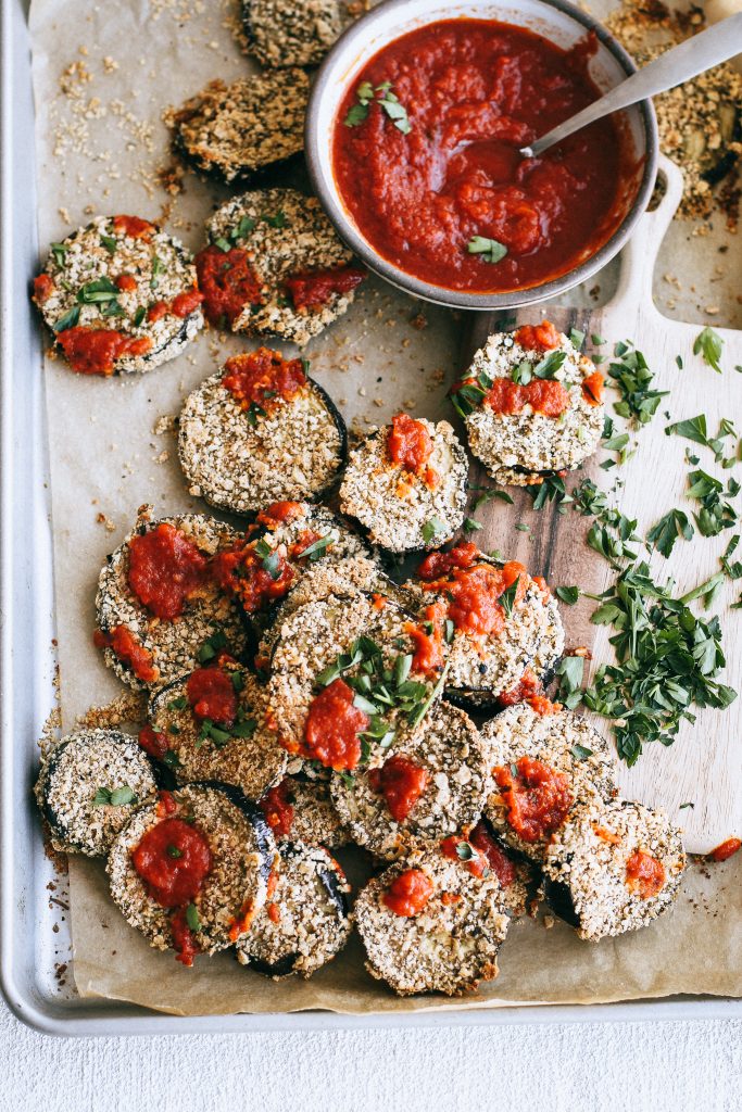 crispy eggplant slices on a baking tray with bowl of marinara and herbs on wood board