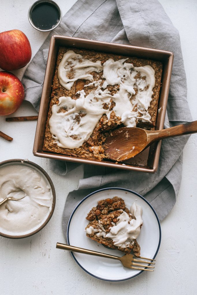 baked apple oatmeal in baking dish with cashew frosting and a slice of baked oatmeal on a white plate with a bowl of cashew frosting
