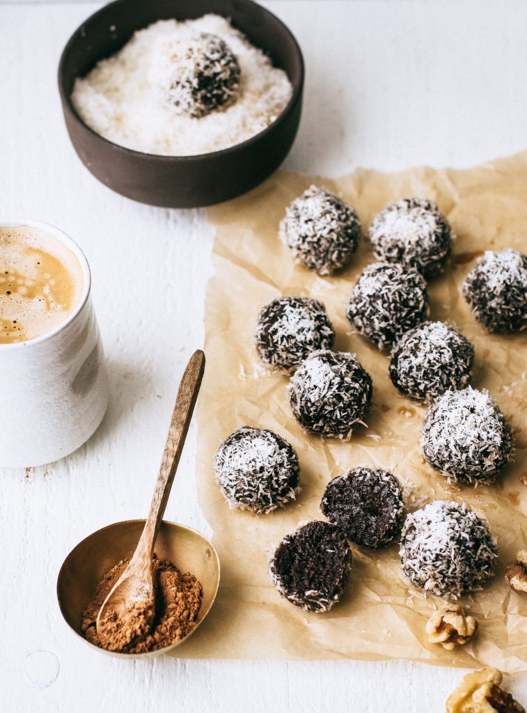 chocolate energy balls rolled in coconut on parchment paper with small bowl and wood spoon and cup of coffee