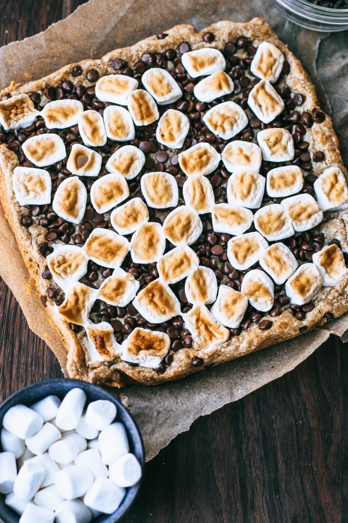 smores bars on white tray on wood cutting board with bowl of marshmallows