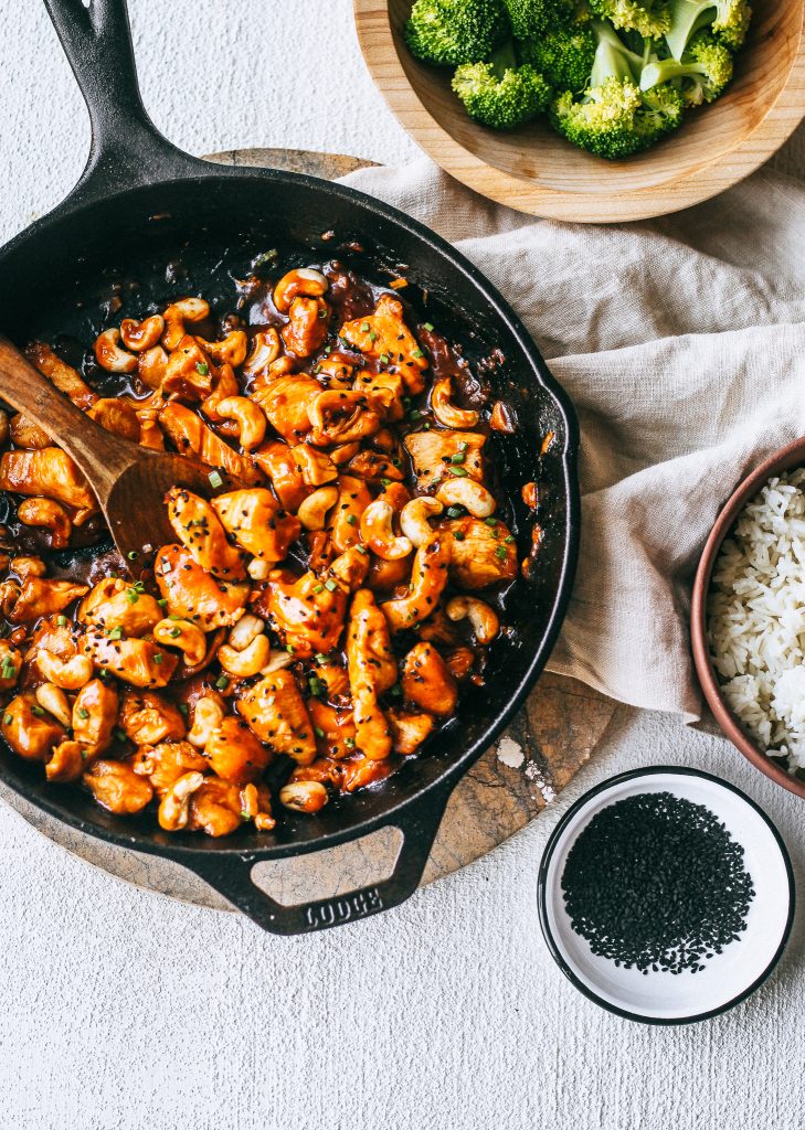 orange cashew chicken in black pan with black sesame seeds in a small white bowl