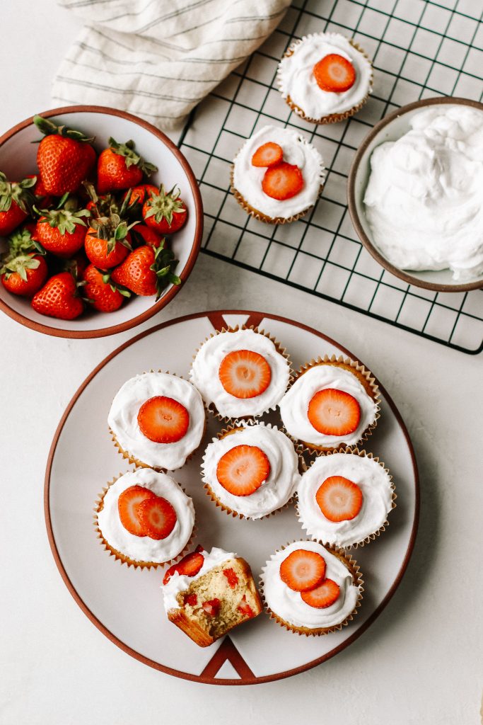 strawberry shortcake cupcakes on a plate with a cooling rack bowl of strawberries and bowl of cool whip
