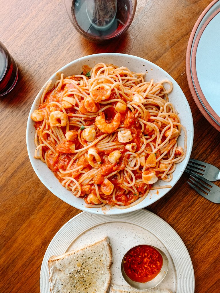 seafood pasta in a bowl with glass of wine, forks, and plate of garlic bread 