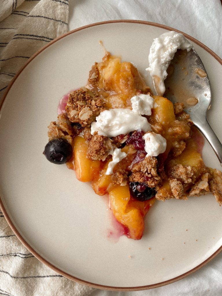 peach and blueberry crisp with a spoon on a plate with cool whip on top