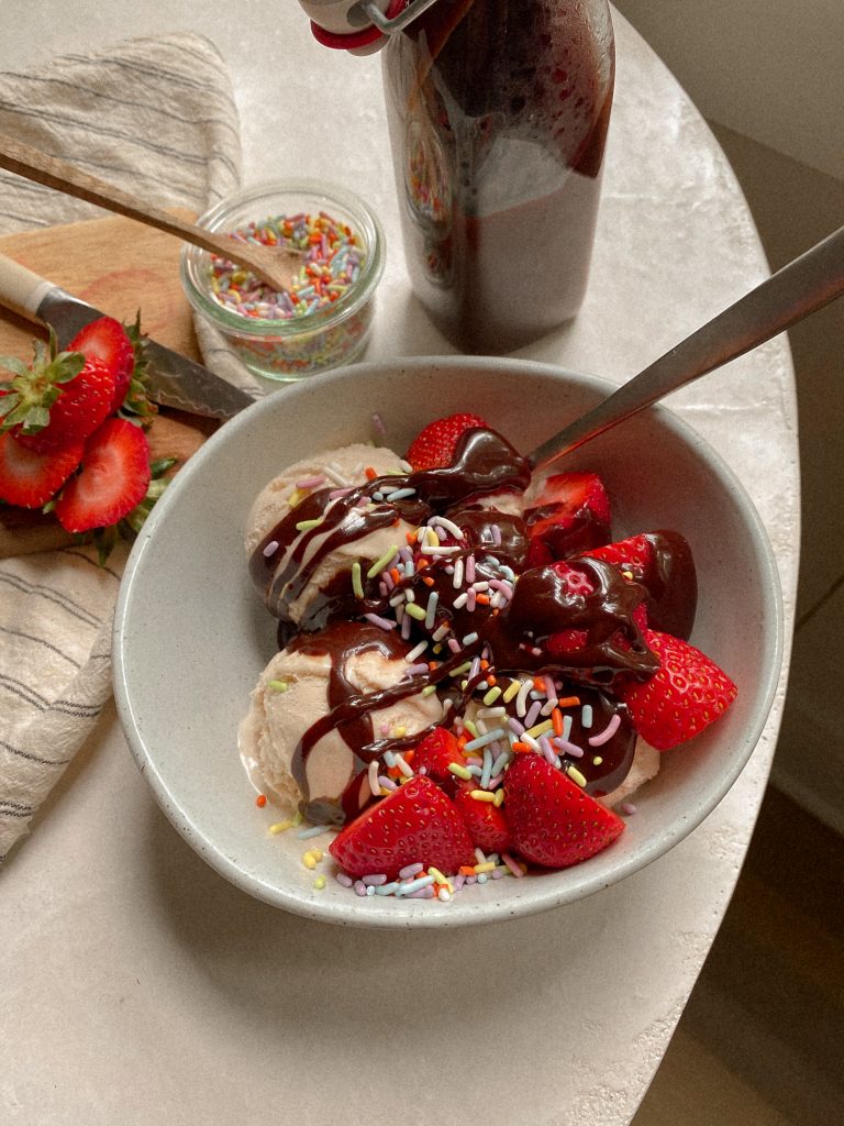 vanilla ice cream in a white bowl with strawberries and hot fudge and sprinkles with small bowl of sprinkles and bottle of hot fudge