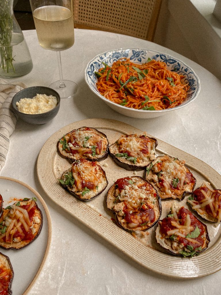 eggplant pizzas on a dish with a bowl of spaghetti and small bowl of parmesan and glass of white wine on a table