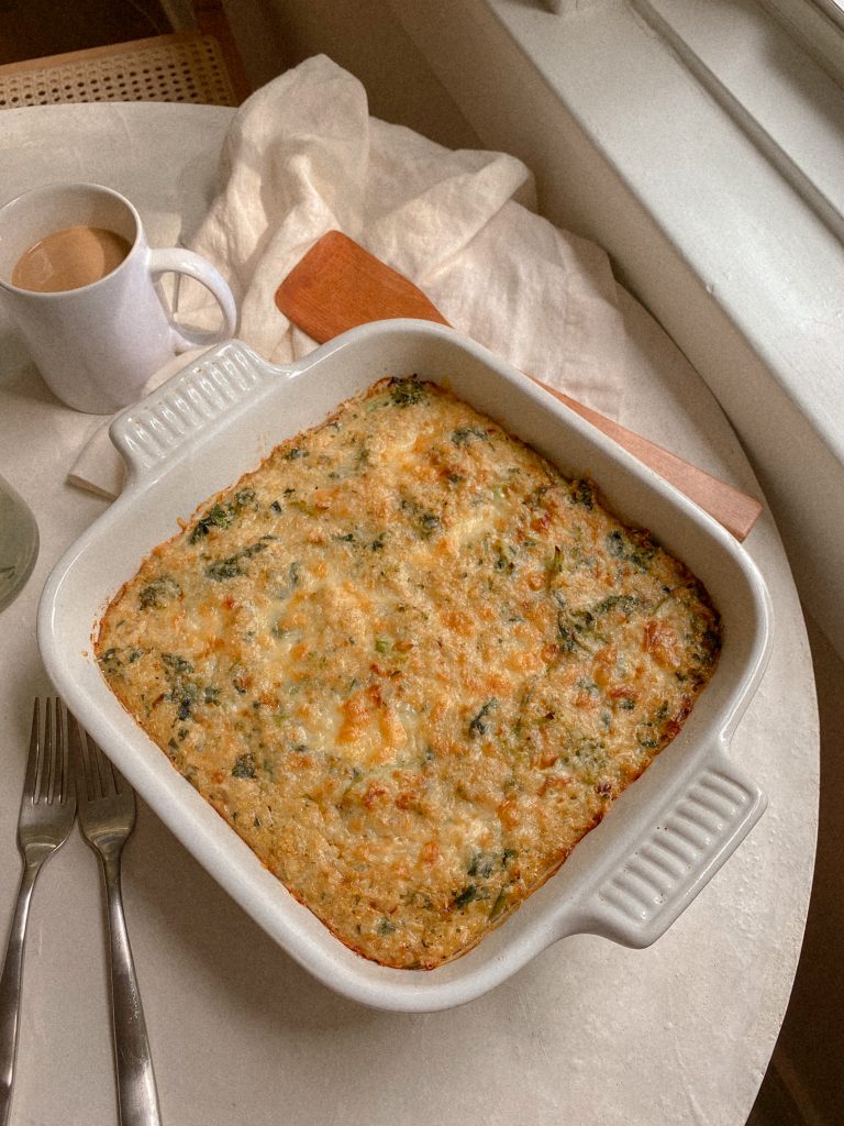 broccoli cheddar quinoa breakfast bake in a white baking dish with a wooden spoon and mug of coffee