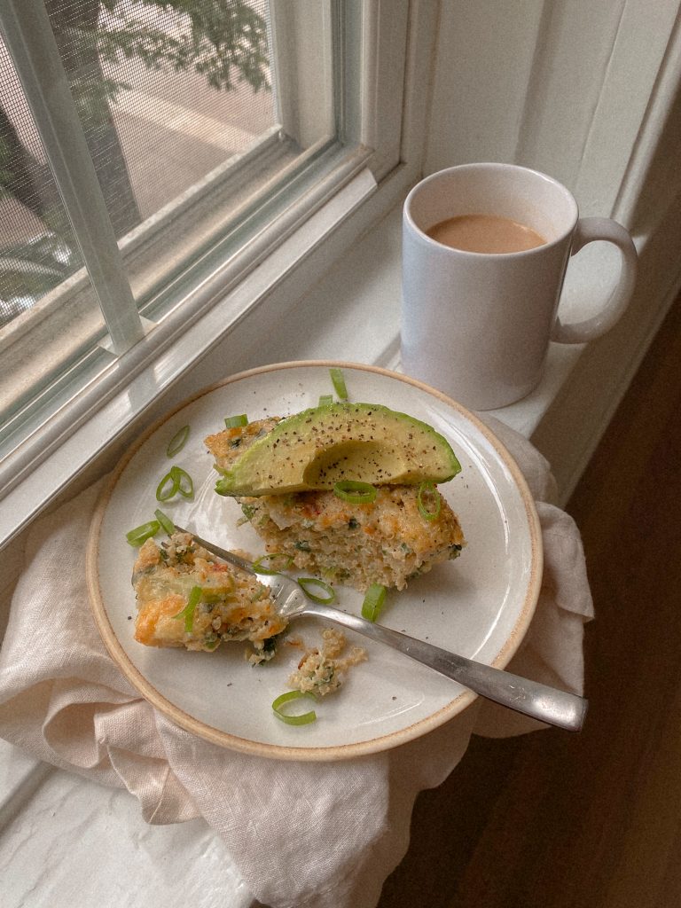 slice of quinoa breakfast bake on a plate with slice of avocado and cup of coffee on a window sill. 