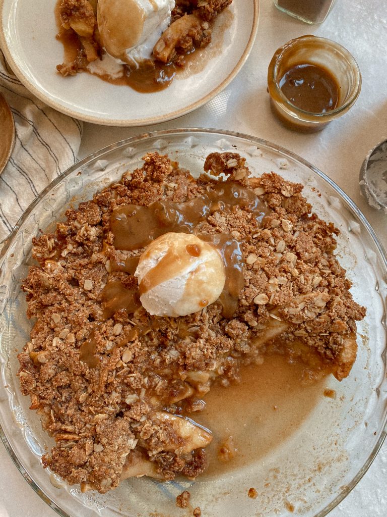 apple crisp with ice cream and caramel in a baking dish with small bowl of caramel and apple crisp slice on a plate