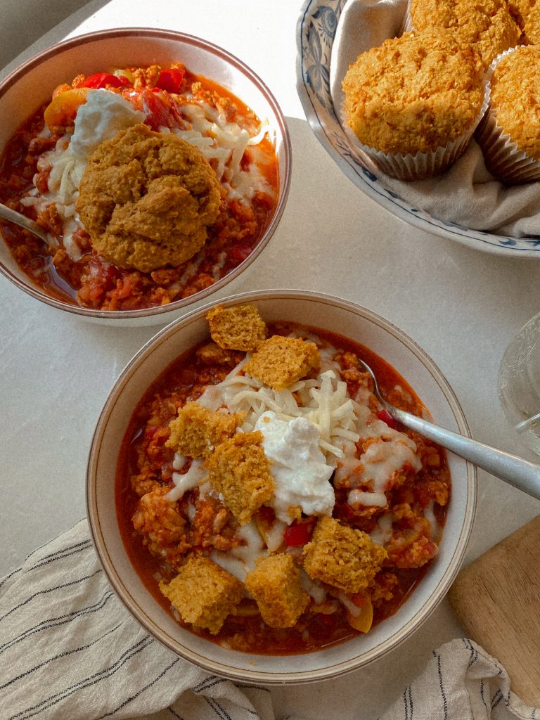 chicken chili in a bowl with cheese and sour cream and cubes of cornbread with a bowl of cornbread muffins