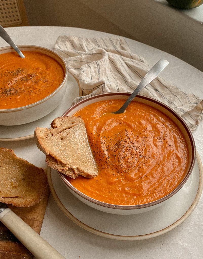 2 bowls of tomato soup with buttered bread and spoons on a table