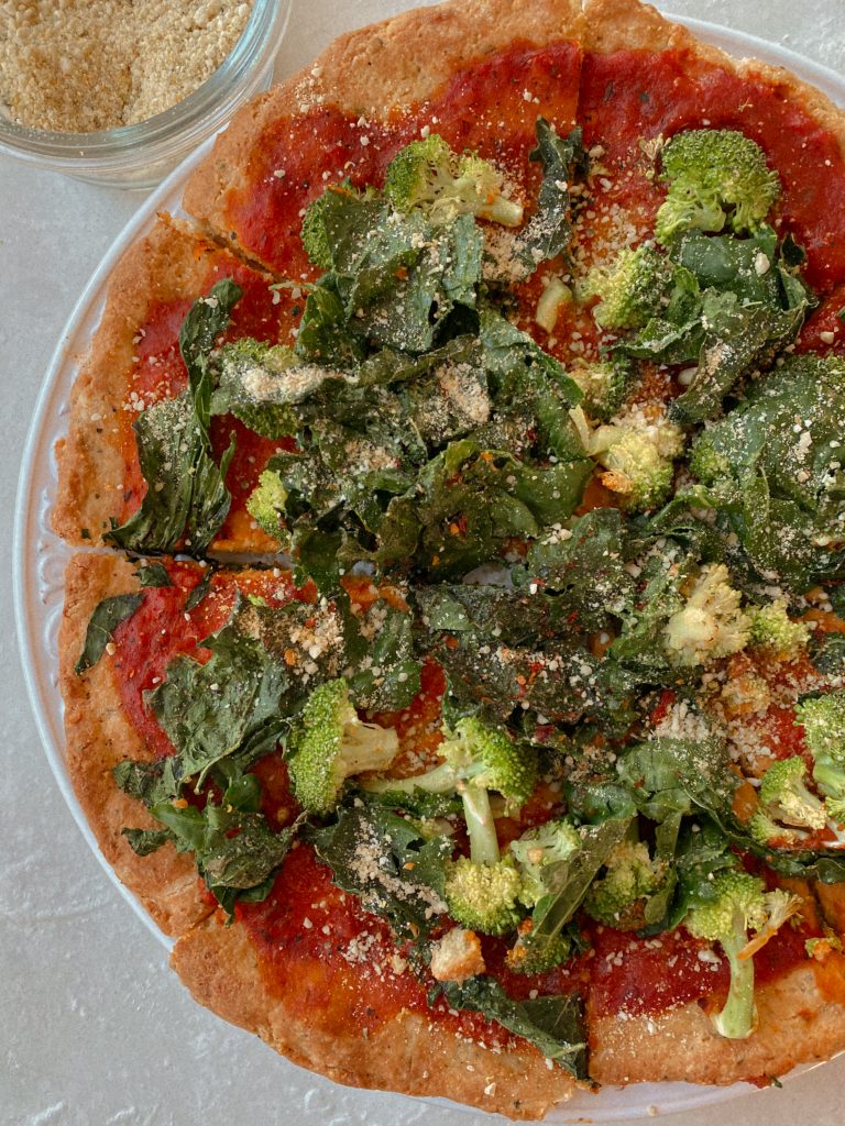 paleo pizza with broccoli and kale on a white plate with small bowl of vegan parmesan