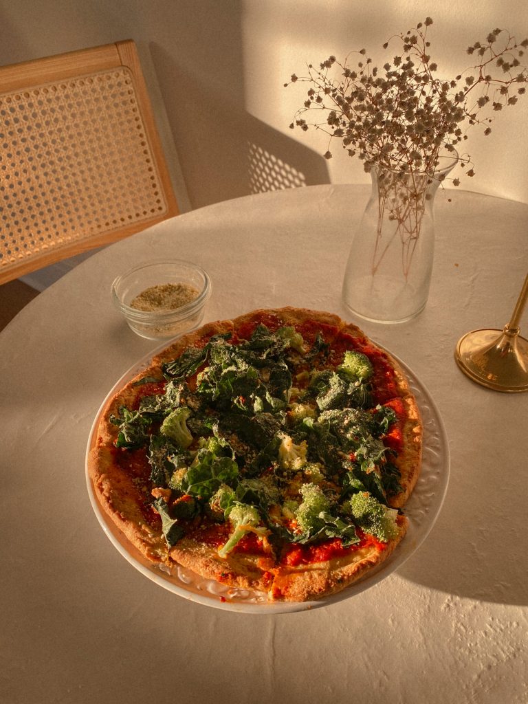 paleo pizza with broccoli and kale on a white plate with small bowl of vegan parmesan and vase with white flowers