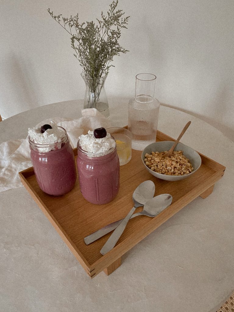 cherry smoothies with whipped cream in jars on a wood tray with bowl of granola, spoons, and jug of water