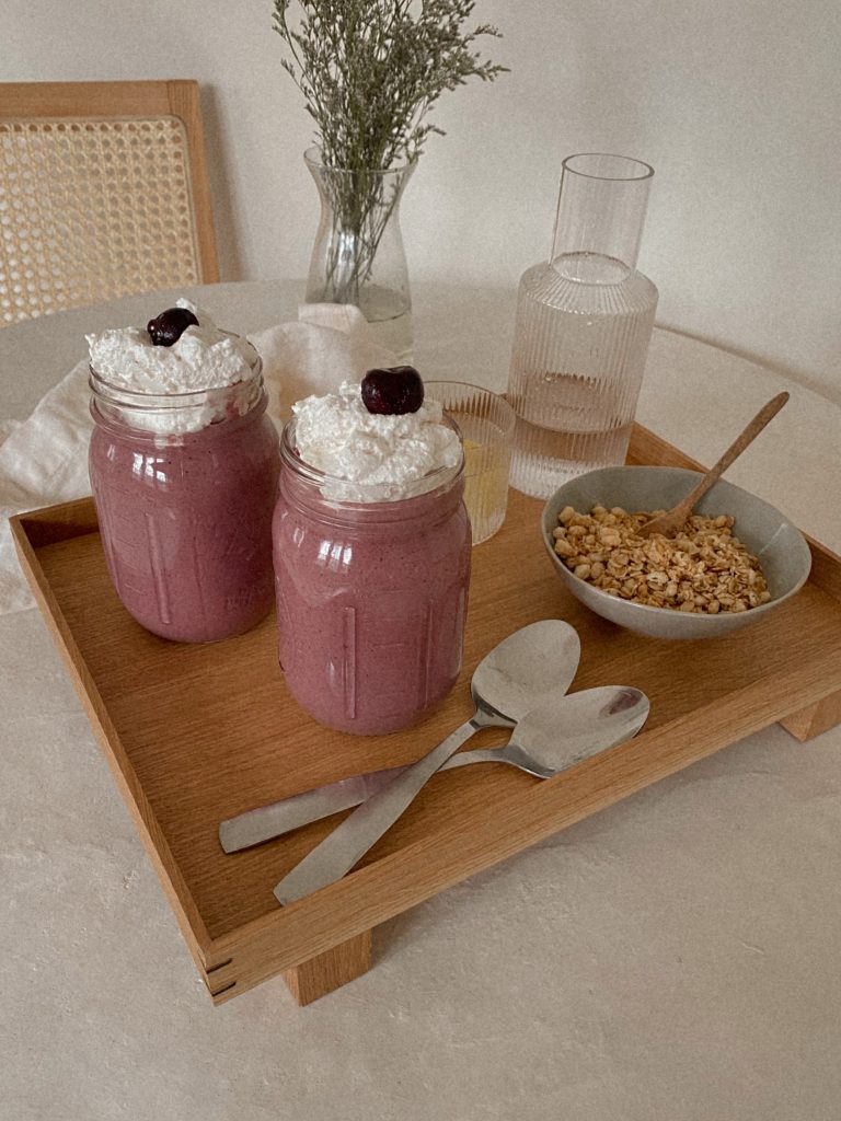 cherry smoothies with whipped cream in jars on a wood tray with bowl of granola, spoons, and jug of water