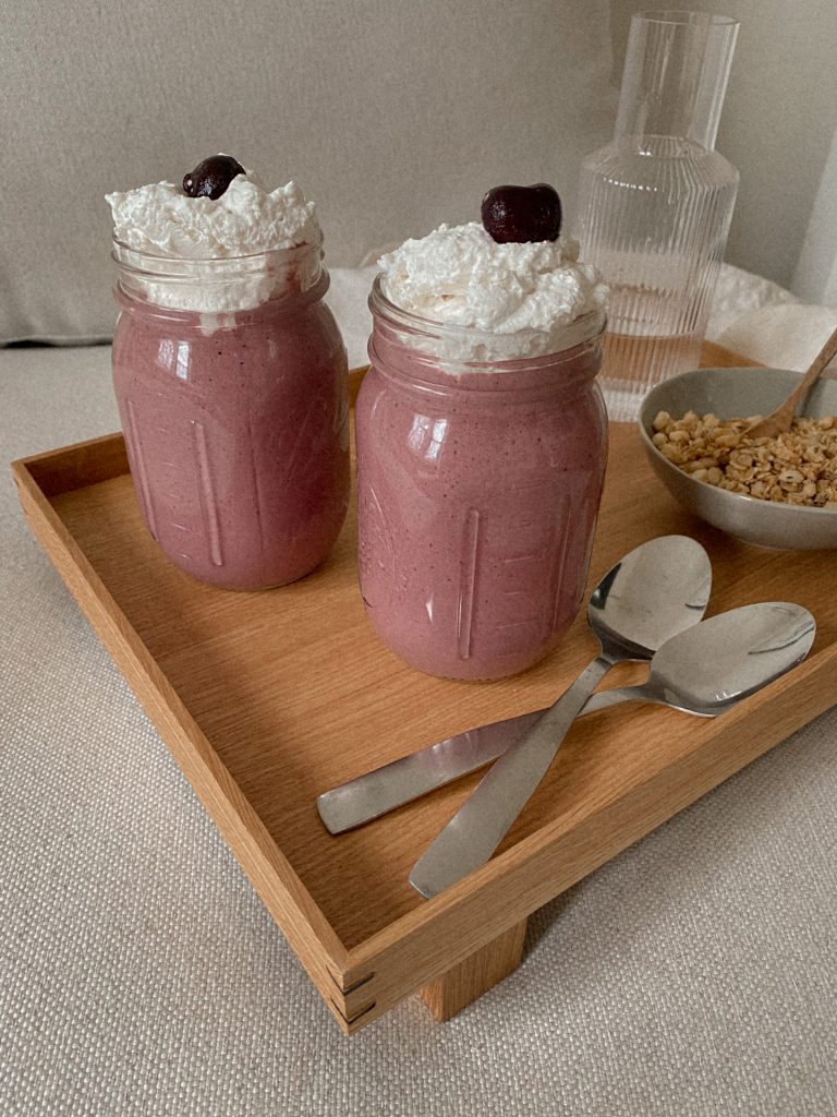 cherry smoothies with whipped cream in jars on a wood tray with bowl of granola, spoons, and jug of water