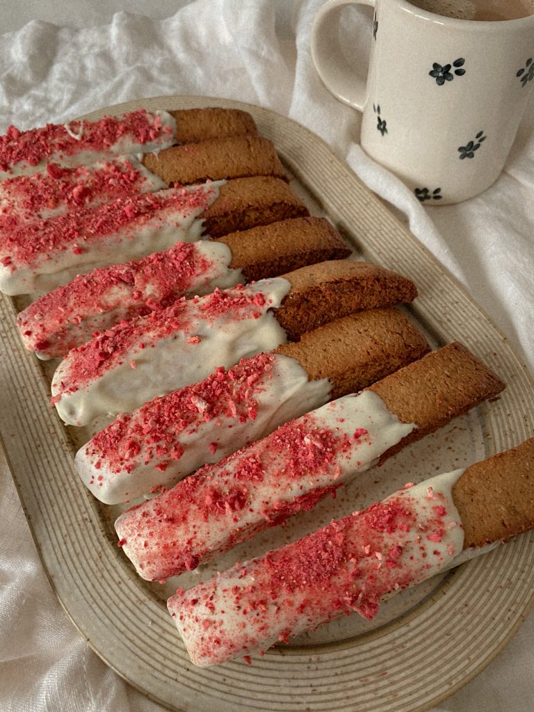 biscotti dipped in white chocolate with crushed freeze dried strawberries on a plate with mug of coffee