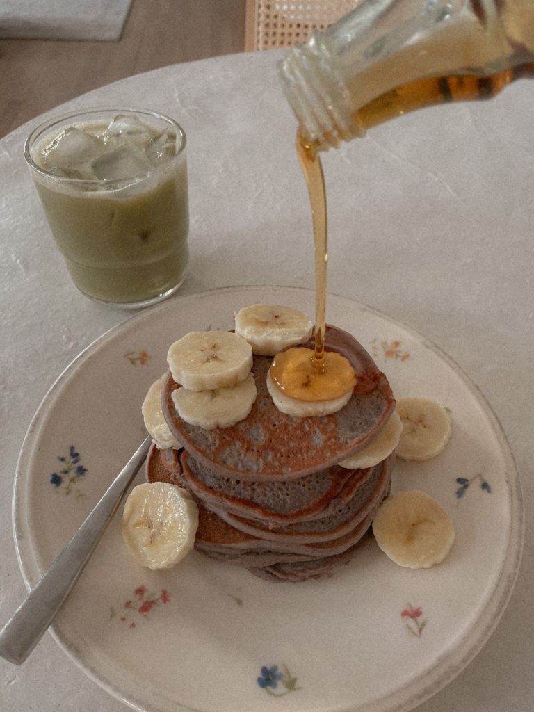 blueberry pancakes with banana slices and maple syrup on a white floral plate with a glass of iced matcha latte