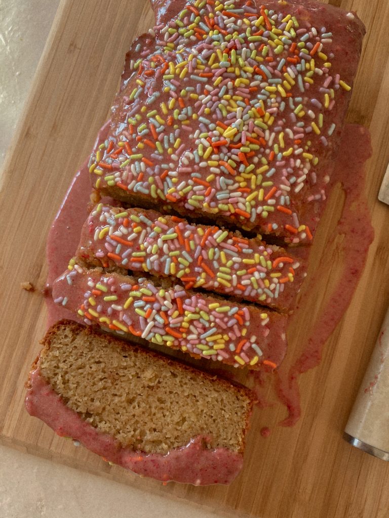 vanilla cake loaf with a pink glaze and sprinkles on a wood cutting board