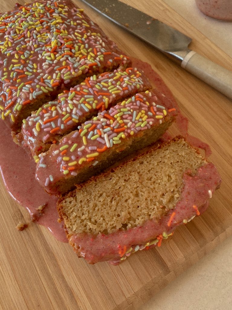 vanilla cake loaf with a pink glaze and sprinkles on a wood cutting board with a knife
