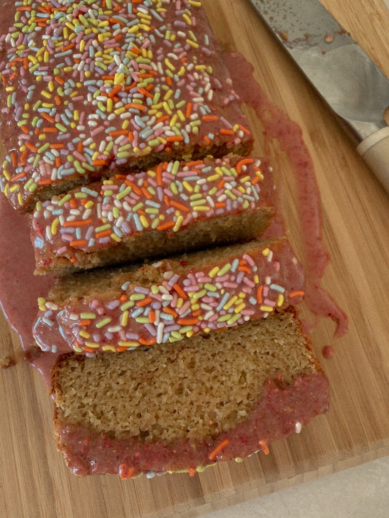 vanilla cake loaf with a pink glaze and sprinkles on a wood cutting board with a knife