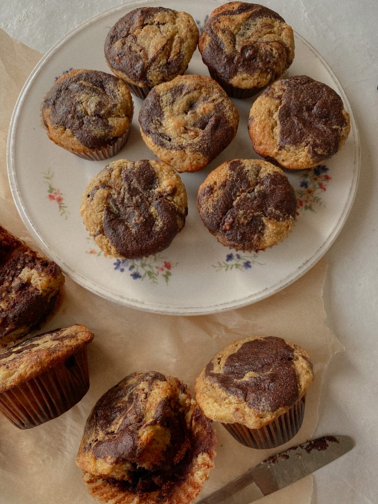 marble chocolate banana muffins on a white floral plate with some muffins on brown parchment paper