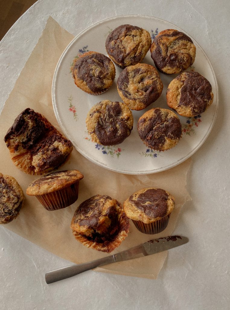 marble chocolate banana muffins on a white floral plate with some muffins on brown parchment paper