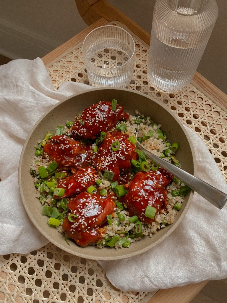 honey sesame chicken and cauliflower rice with green onions in a bowl with a spoon on a cane chair with a white napkin